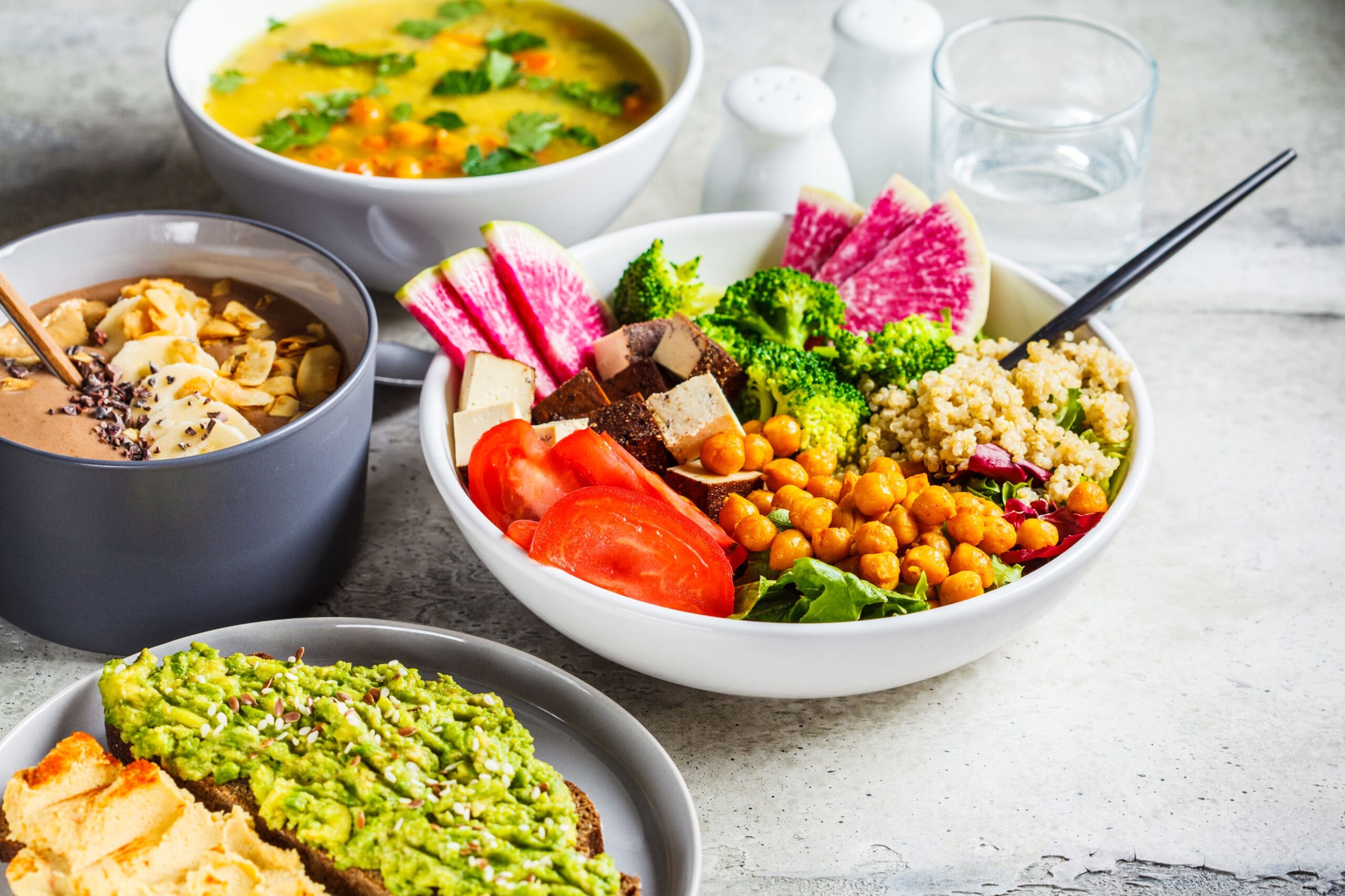 Assorted bowls of colorful plant-based dishes, including a salad bowl with vegetables and chickpeas, avocado toast, and a bowl of soup.