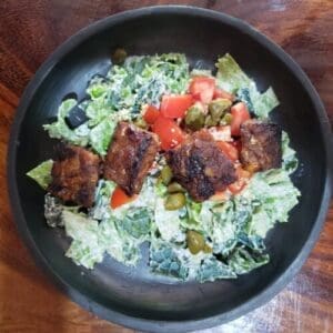 Kale Caesar salad topped with marinated tempeh pieces, tomatoes, and croutons, served in a large black bowl on a hardwood table