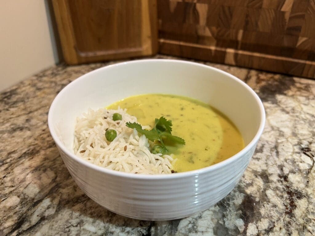 White bowl on a granite countertop filled with yellow yogurt-based kadhi curry and basmati rice with peas, garnished with fresh cilantro leaves.