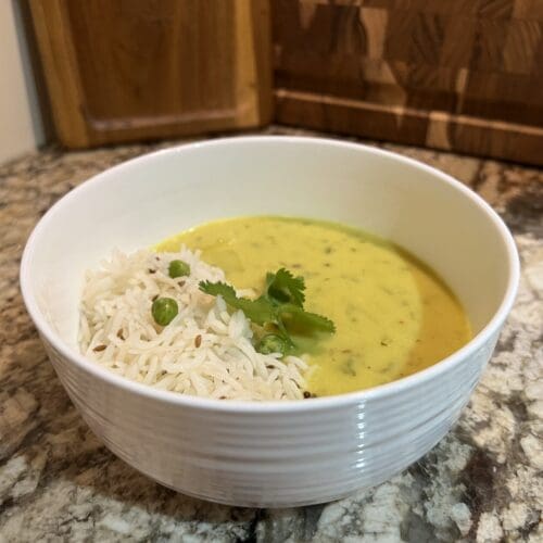 White bowl on a granite countertop filled with yellow yogurt-based kadhi curry and basmati rice with peas, garnished with fresh cilantro leaves.