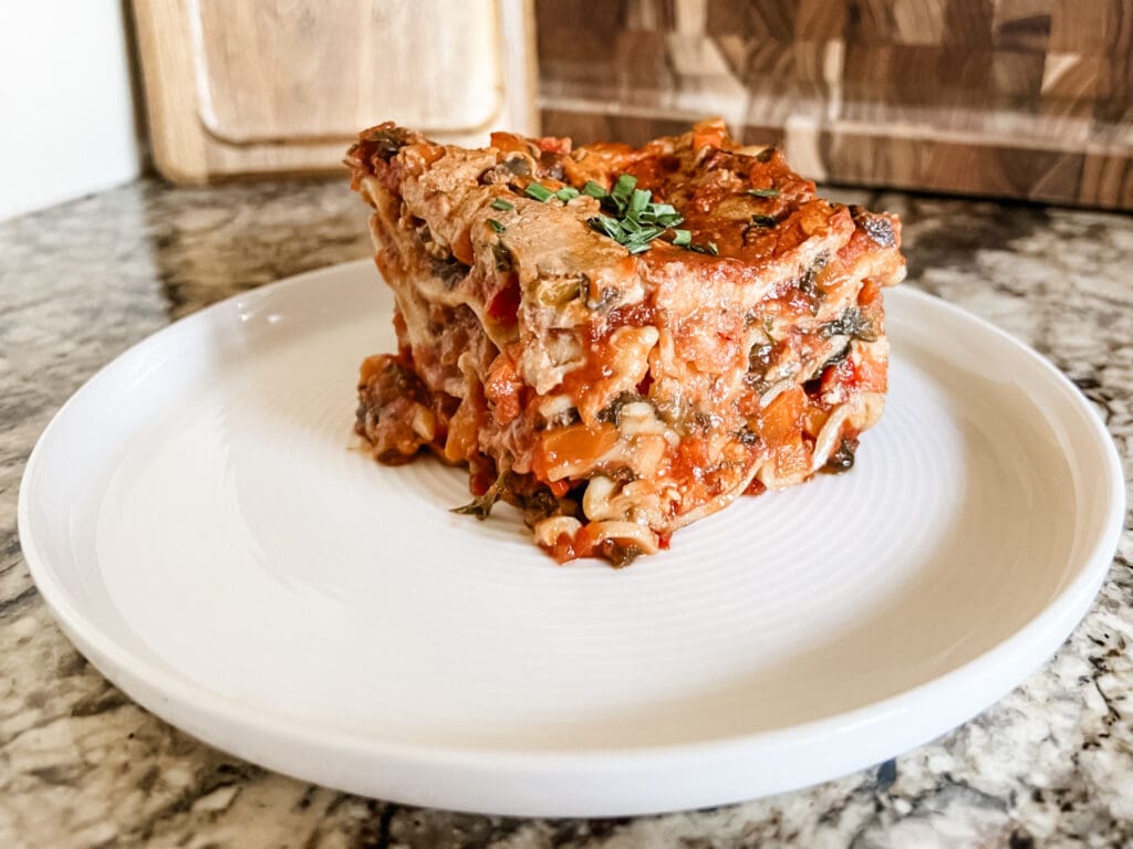 A close-up of a slice of hearty lasagna on a white plate. The lasagna has multiple layers of pasta, vegetables, and a rich tomato sauce, topped with melted cheese. The filling includes a mix of red and green vegetables, giving it a colorful and appetizing appearance. The plate sits on a marble countertop, and the lasagna is garnished with fresh herbs on top.