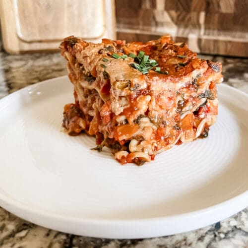 A close-up of a slice of hearty lasagna on a white plate. The lasagna has multiple layers of pasta, vegetables, and a rich tomato sauce, topped with melted cheese. The filling includes a mix of red and green vegetables, giving it a colorful and appetizing appearance. The plate sits on a marble countertop, and the lasagna is garnished with fresh herbs on top.