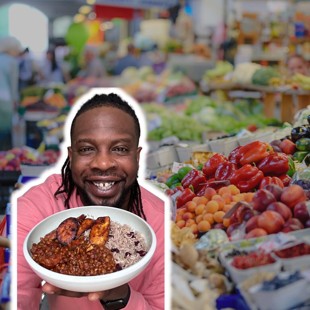 Ebenezer Odeniyi, a Black man with long locs and a nose ring, smiling and holding a bowl of plant-based food (rice, beans, and plantains). He wears a pink sweatshirt in front of a colorful market filled with fresh produce, symbolizing the vibrancy of plant-based eating.