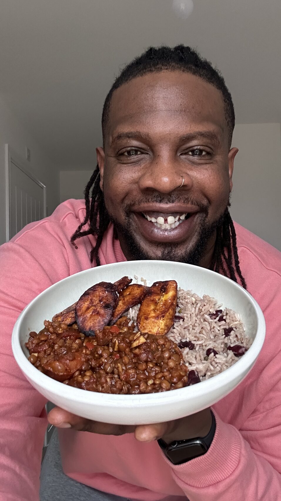 Ebenezer Odeniyi is in a pink shirt, smiling while holding a bowl of rice and beans, seasoned lentils, and crispy fried plantains. He is showcasing a colorful, plant-based meal.