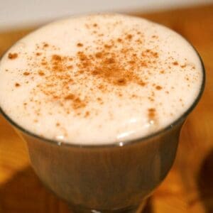 A closeup shot of a glass of apple postre, a mix of merangue and shredded apple, topped with powdered cinnamon, on a wood table.